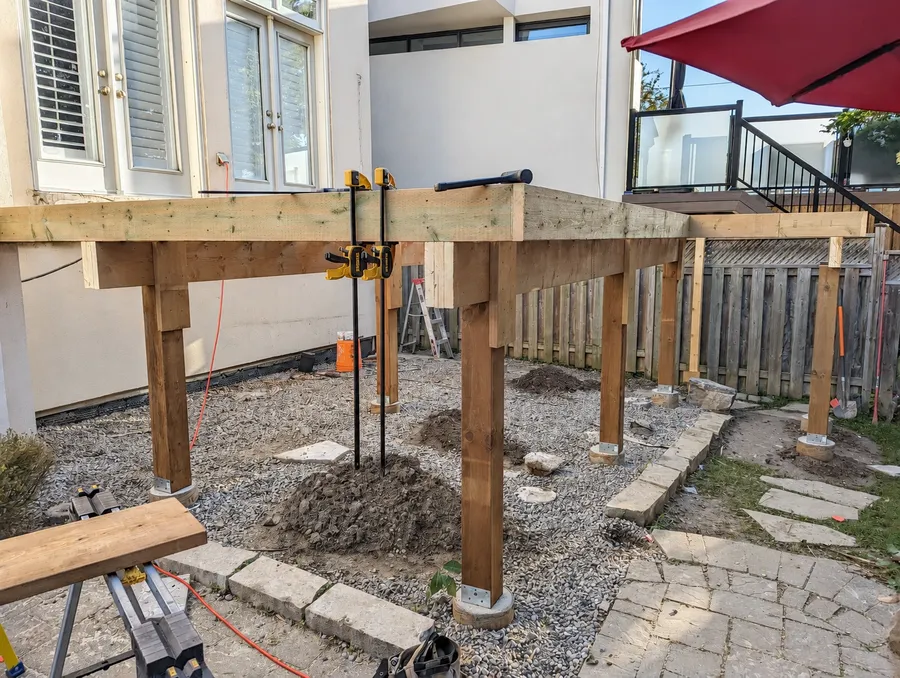Deck frame under construction beside a house, with timber posts on concrete piers and perimeter framing clamped in place
