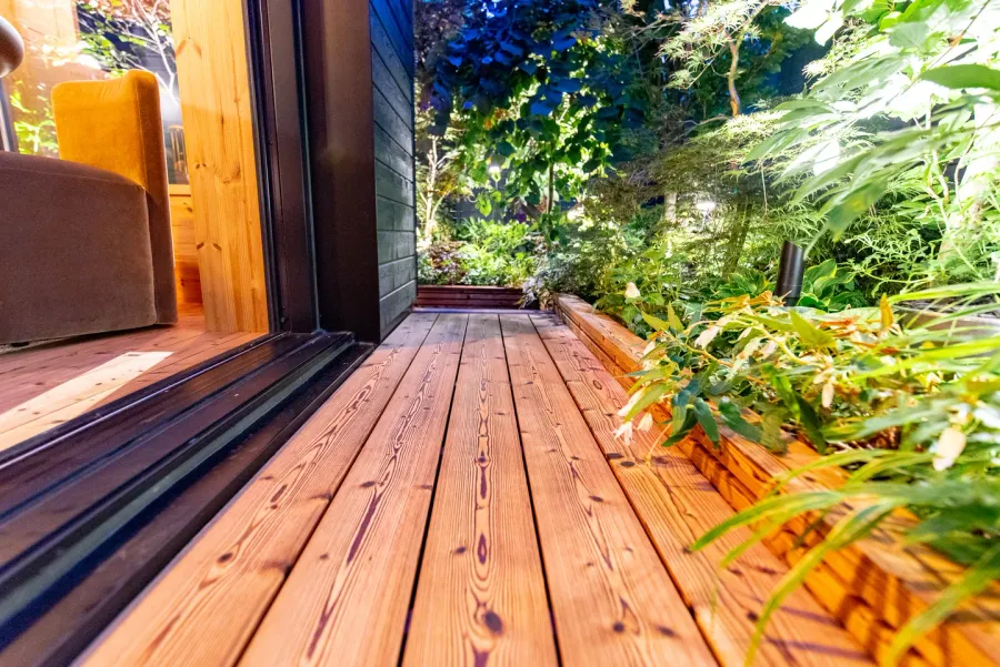 Close view of knotted wood decking alongside an open garden-room doorway and leafy planting.
